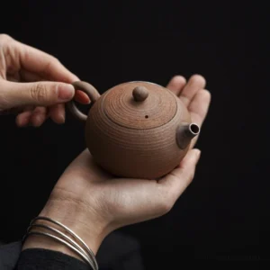 Mini handmade rough clay teapot held in hand showing ergonomic handle design